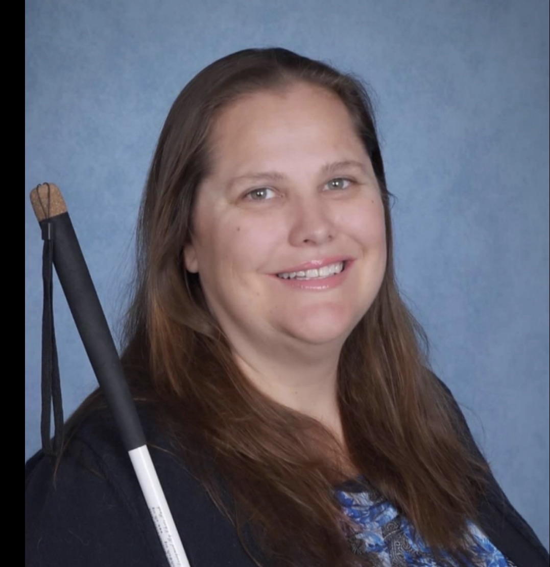 Laura Millar, a white woman with long brown hair, smiling in a professional portrait with a white cane visible beside her against a soft blue background.